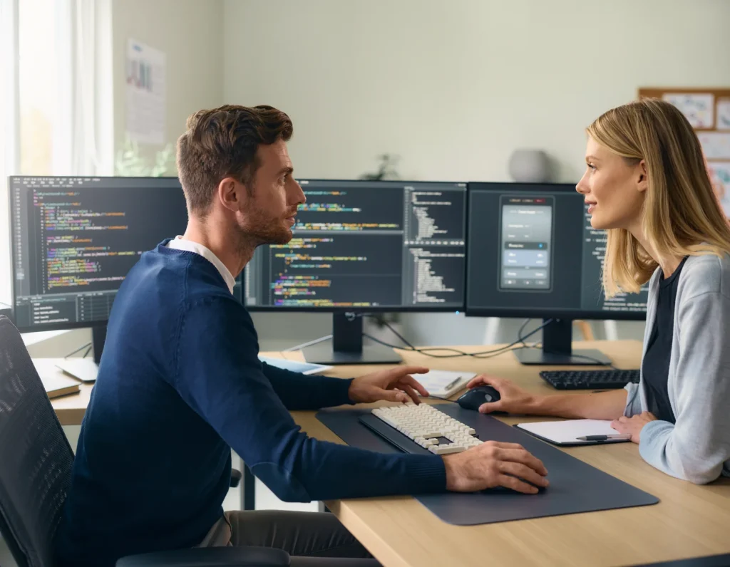 Zwei Personen besprechen Software an einem Arbeitsplatz mit mehreren Monitoren.